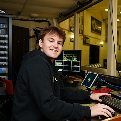 A young man sitting a studio mixing board. He smiles at the camera.